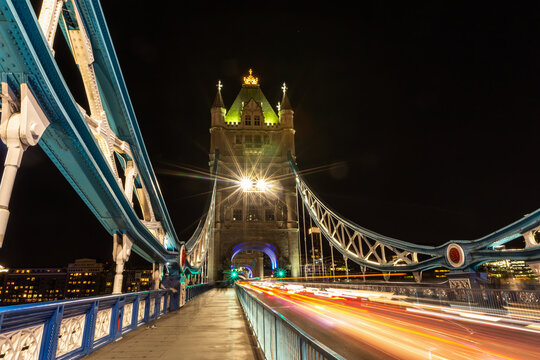 View of Tower Bridge at night with light trails from passing traffic on the road, illuminated gothic towers, and blue steel suspension structure in London, England, United Kingdom.