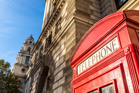 View of a classic red telephone box standing in front of a historic stone building under a clear blue sky in London, England, United Kingdom.