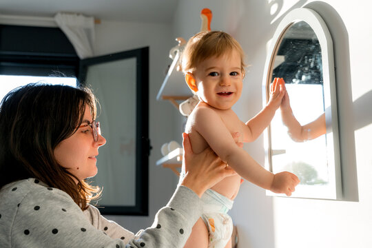 Mother lifting baby by window in bright home nursery light