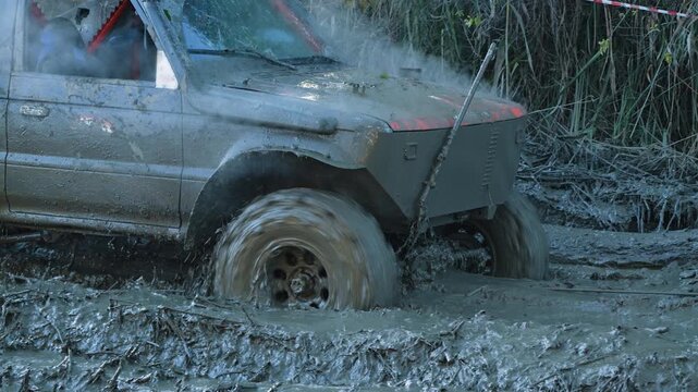 Modified 4x4 vehicle struggles in thick, deep mud. Spinning wheels splash dirt water during extreme off roading competition. Steam rises from overheating engine. Rugged, powerful, dirty scene