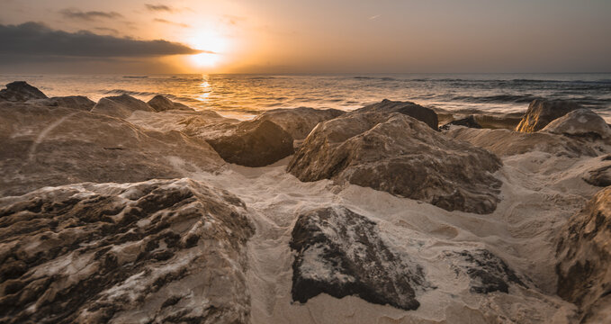 View of large rocks on a sandy beach at sunset with ocean waves rolling in under a golden sky in Gulf Shores, Alabama, United States.