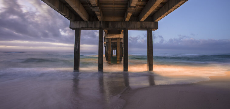 View of the underside of a pier at sunset with soft waves washing onto the sandy beach in Gulf Shores, Alabama, United States.