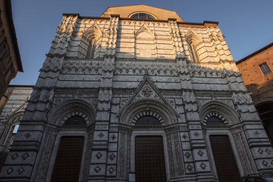 View of the ornate white and dark green marble facade of the Siena Cathedral illuminated by warm golden hour light under a clear blue sky in Siena, Tuscany, Italy.