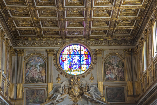 View of the ornate interior of a historic church featuring a vibrant circular stained glass window, intricate gold-leaf coffered ceiling, and detailed religious frescoes in Rome, Lazio, Italy.