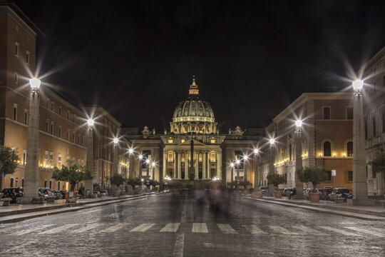 View of St. Peter's Basilica illuminated at night with glowing street lamps and a cobblestone street in Rome, Lazio, Italy.
