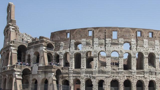 View of the Colosseum in Rome, showing its iconic ancient stone arches and weathered facade under a clear blue sky with tourists visible on the upper levels, Roma, Lazio, Italy.