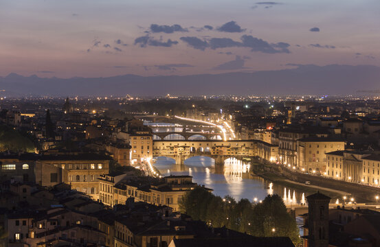 View of the historic Ponte Vecchio bridge spanning the Arno River at twilight with illuminated buildings and city lights in Florence, Tuscany, Italy.