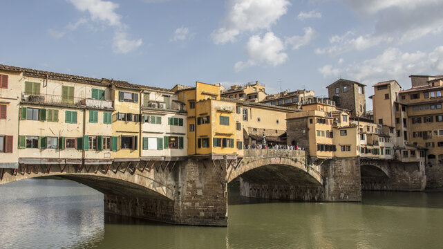 View of Ponte Vecchio bridge over the Arno River in Florence, featuring historic buildings with green shutters, stone arches, and people walking on the bridge. Florence, Tuscany, Italy.