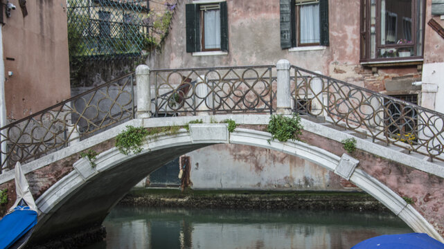 View of a stone arch bridge over a canal with ornate metal railings and weathered building facades in the background in Venice, Veneto, Italy.