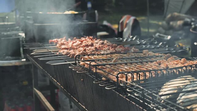 Skewers of meat are being grilled outdoors at a gathering. Smoke rises from the grill as people enjoy the sunny atmosphere