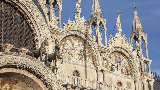 View of the ornate facade of St. Mark's Basilica featuring intricate golden mosaics, bronze statues of horses, and detailed marble carvings in Venice, Veneto, Italy.