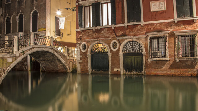 View of a quiet canal in Venice at night, featuring historic brick buildings, a stone bridge reflecting in the calm water, and warm street lighting in Venice, Veneto, Italy.