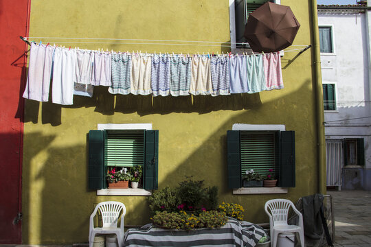 View of laundry hanging on a line against a vibrant yellow wall with green shutters and potted flowers in Burano, Venice, Veneto, Italy.