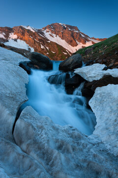 View of Noori lake with a stream of meltwater flowing through snowbanks, framed by rocky terrain and snow-capped mountains under a clear blue sky in Noori Top, Azad Kashmir, Pakistan.