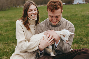 Obraz premium Couple in cozy sweaters cuddle a gentle goat kid in a grassy field, sharing warmth and smiles, a candid countryside moment of affection and relaxed lifestyle.