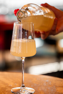 Hand being poured a cloudy fermented drink from a flip-top glass bottle into an elegant stemware glass on a wooden table, close-up view.