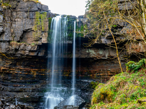 View of Ashgill Force waterfall cascading over a rugged rocky cliff face surrounded by mossy stones and bare tree branches in Alston, England, United Kingdom.