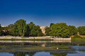 Fototapeta premium Blick über das Elstermühlgraben- oder Elsterflutbett-Ufer mit Bäumen und historischen Gebäuden im Sonnenschein im Sommer, Leipzig, Sachsen, Deutschland