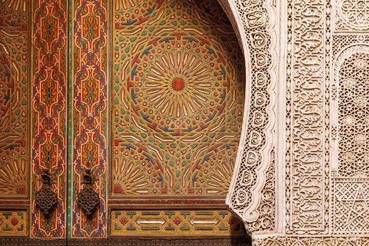 Close-up of an ornately painted wooden door panel beside an intricately carved stucco archway at the Mausoleum of Sidi Ahmed al-Tijani in the Fez medina, Morocco, a UNESCO World Heritage Site.