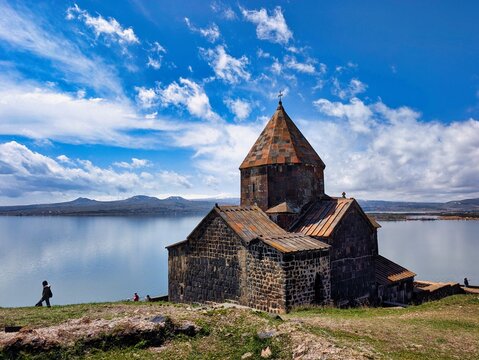 View of Sevanavank monastery overlooking Lake Sevan under a bright blue sky with clouds, with a person walking on the grassy hillside in Sevan, Gegharkunik Province, Armenia.