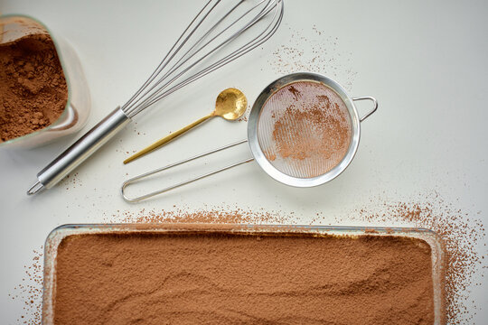 Tiramisu dessert with cocoa powder in glass dish, with sieve, whisk, spoon and cocoa on table