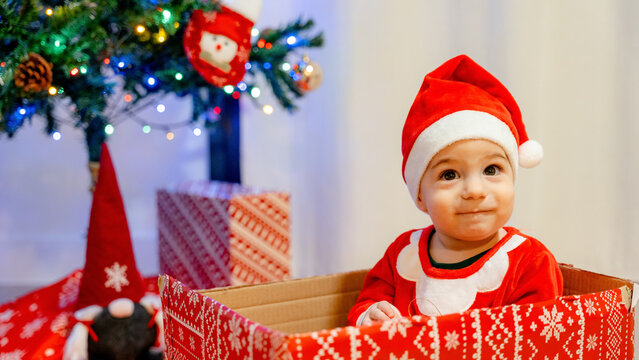 Baby in Santa hat sitting in gift box by Christmas tree