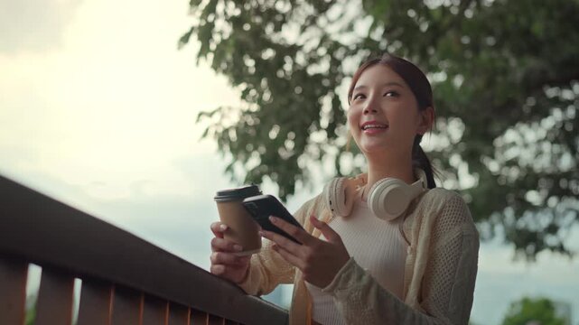 Happy Asian woman holding coffee cup and using smartphone at city park balcony during sunset, cheerful female wearing headphones relaxed online texting for urban digital lifestyle and mental wellness