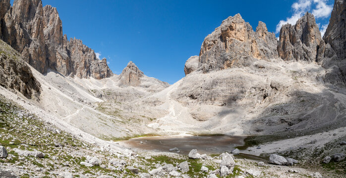 View of the Dolomites mountain range with rocky peaks, a small alpine lake, and sparse green vegetation in San Martino di Castrozza, Trentino-Alto Adige, Italy.