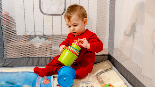 Toddler in red pajamas stacking cups in playpen at home