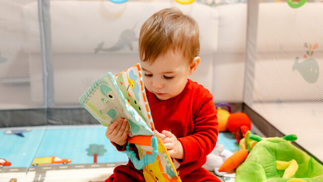 Baby reading soft cloth book in playpen with toys