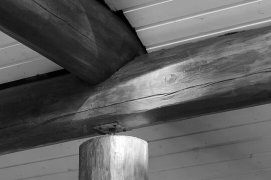 Close-up of a rustic log beam ceiling and round support post with visible timber grain, knots and metal bracket. Monochrome interior detail emphasizing texture, craftsmanship, structural stability 