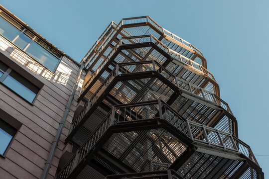 Low-angle view of a metal fire escape staircase attached to a modern building facade, showing geometric lines, urban architecture and industrial textures against a clear blue sky