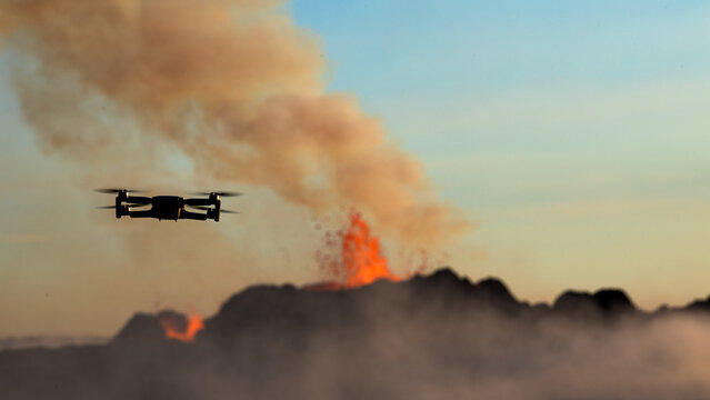 View of a drone flying in front of a volcanic eruption with smoke and glowing lava in the background in Iceland.