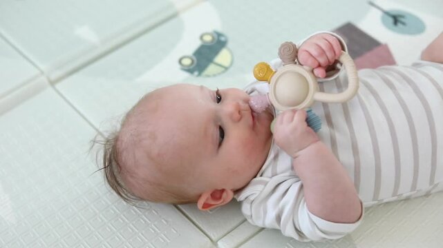 Close-up portrait of a baby chewing a silicone teething toy while lying on a soft blanket, soothing sore gums during teething in a bright nursery setting.
