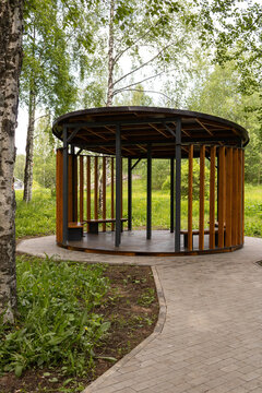 Modern circular wooden pavilion gazebo in a city park surrounded by birch trees and green grass in early summer. Urban park landscape design concept.