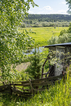 Old rustic wooden shed and broken fence by a small river and green meadow field in the Far North of Russia. Scenic summer northern landscape.