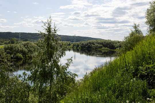 Scenic view of a calm river reflecting sky surrounded by green trees and grassy fields in the Far North of Russia. Natural subarctic summer landscape.