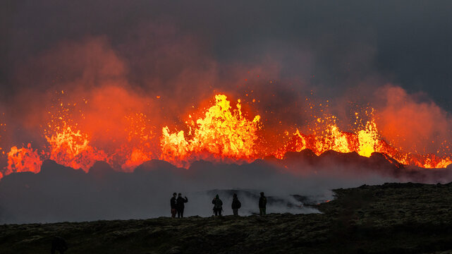 View of a dramatic volcanic eruption with glowing lava fountains erupting against a dark sky, with people standing in the foreground watching the natural phenomenon in Iceland.