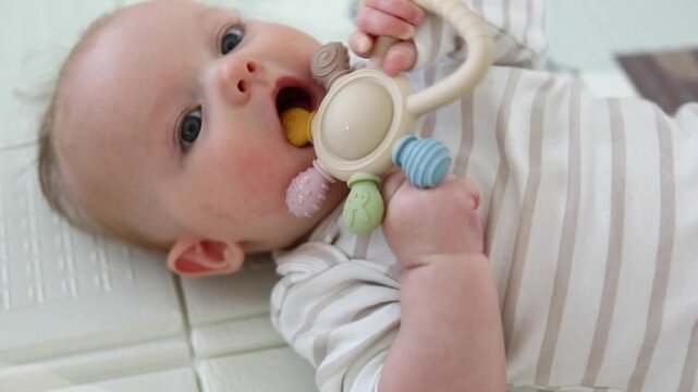 Close-up portrait of a baby chewing a silicone teething toy while lying on a soft blanket, soothing sore gums during teething in a bright nursery setting.