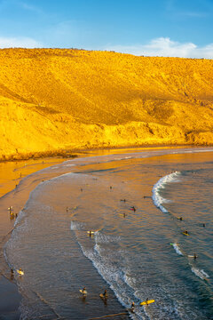 View of Imsouane beach with surfers riding waves in the Atlantic Ocean under golden hour light against dramatic cliffs in Imsouane, Souss-Massa, Morocco.
