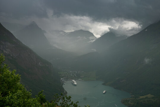 View of the Geirangerfjord with cruise ships docked in the water, surrounded by steep mountains and lush green slopes under a dramatic cloudy sky in Geiranger, More og Romsdal, Norway.