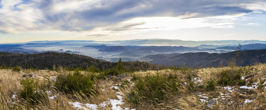 View of the Grazer Bergland mountain range with dry grass, scattered snow, and distant valleys under a cloudy sky in Graz, Styria, Austria.