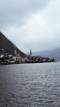 Cinematic pan of Hallstatt village and lake with falling snow. Scenic Austrian alpine village during gentle winter snowfall. Panning shot of iconic Hallstatt viewpoint, vertical resolution.