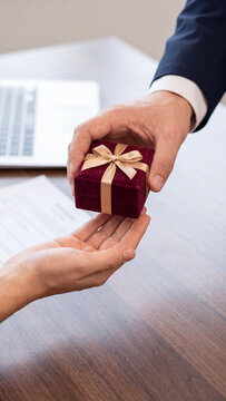 A close-up of a male businessman hand giving a small red velvet jewelry gift box with a gold ribbon, to a colleague at a wooden office desk, with a laptop in the background.