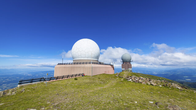 View of the Koralpe radar station with its large white radome and a smaller secondary dome situated on a grassy mountain peak under a clear blue sky in Weinebene, Styria, Austria.