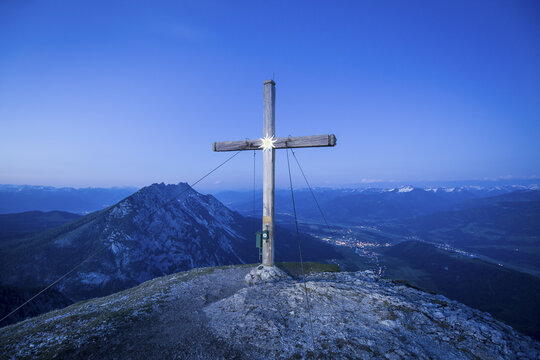 View of the summit cross on Stoderzinken at twilight with the illuminated valley of Grobming visible in the distance. Grobming, Steiermark, Austria.