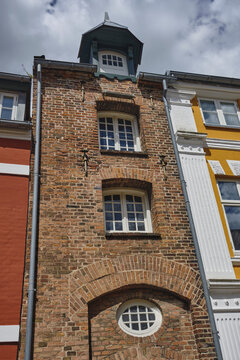View of a small masonry warehouse with an ornamental dormer and hoisting gear, featuring a red brick facade, white-framed windows, and a round window under a brick arch in Ribe, Denmark.