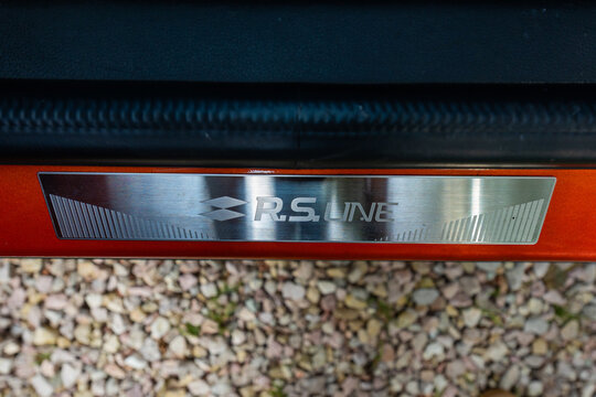 Stainless door sill insert engraved with R.S. LINE and Renault double diamond rests at the threshold against metallic orange paint over pebbles in soft daylight.