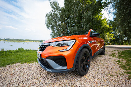 Orange Renault Arkana RS Line parks on a gravel path by a lake with trees. Low wide angle view highlights aggressive bumper, C shaped LED DRLs, gloss black wheels, and RS Line badging.