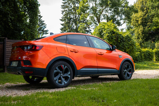 A vivid orange Renault Arkana RS Line parks on a gravel path beside a lawn, rear three quarter view shows sloping roofline, gloss black trim, dual exhaust, and alloy wheels.